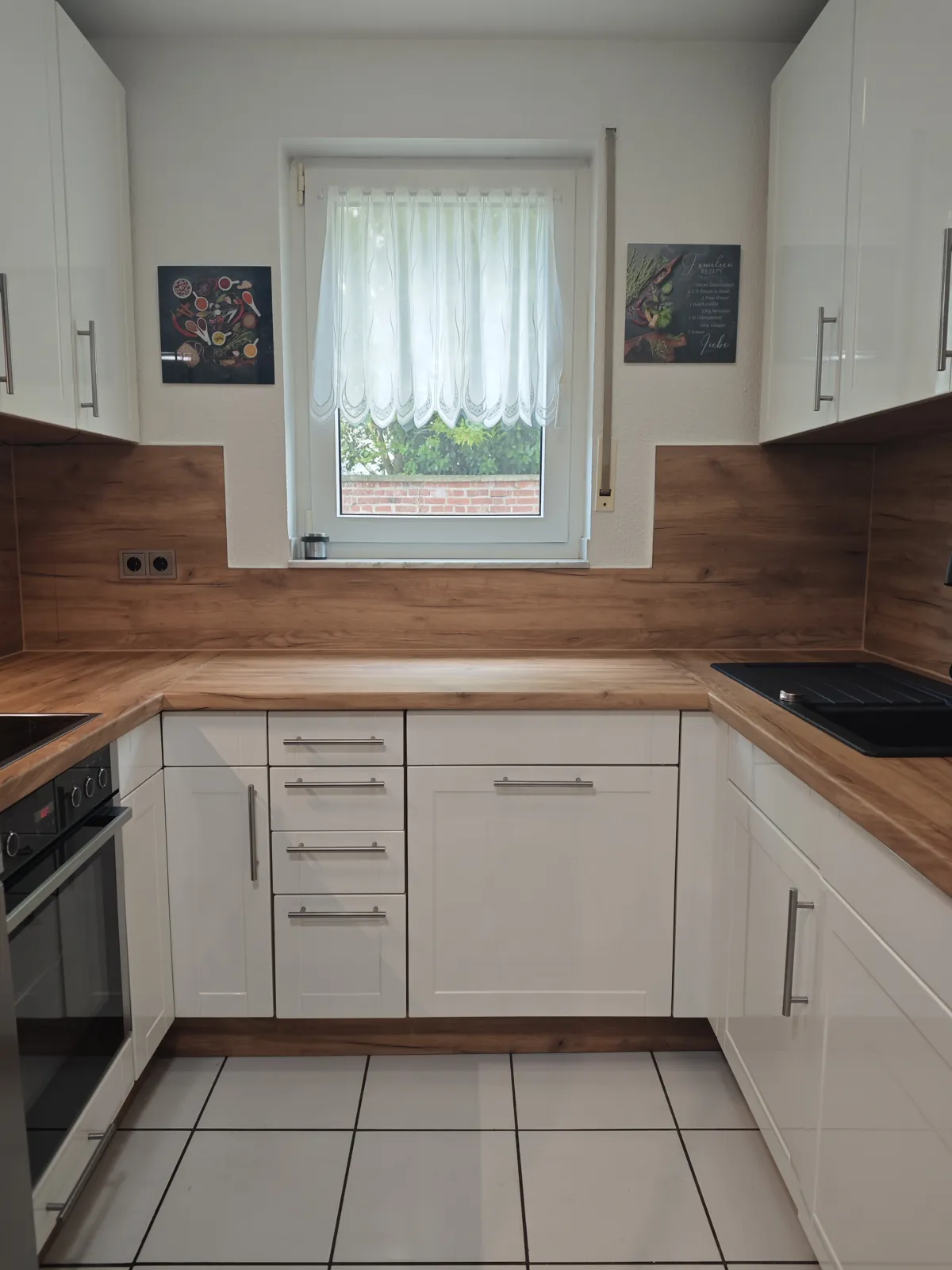 U-Küche in Köln nach der Folierung: Fronten in Weiß hochglanz mit Rahmenoptik und Edelstahlgriffen, Arbeitsplatte und Küchenrückwand in warmer Eichenoptik, Fenster mit Gardine