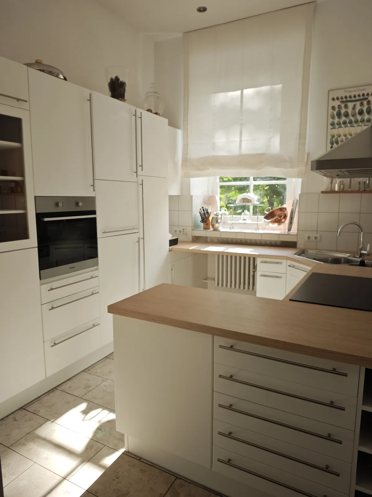 Große Küche mit weißen Fronten und Stangengriffen: Hochschrankwand mit Backofen, Halbinsel mit Arbeitsplatte in heller Eichenoptik und Kochfeld, Fenster mit Raffrollo