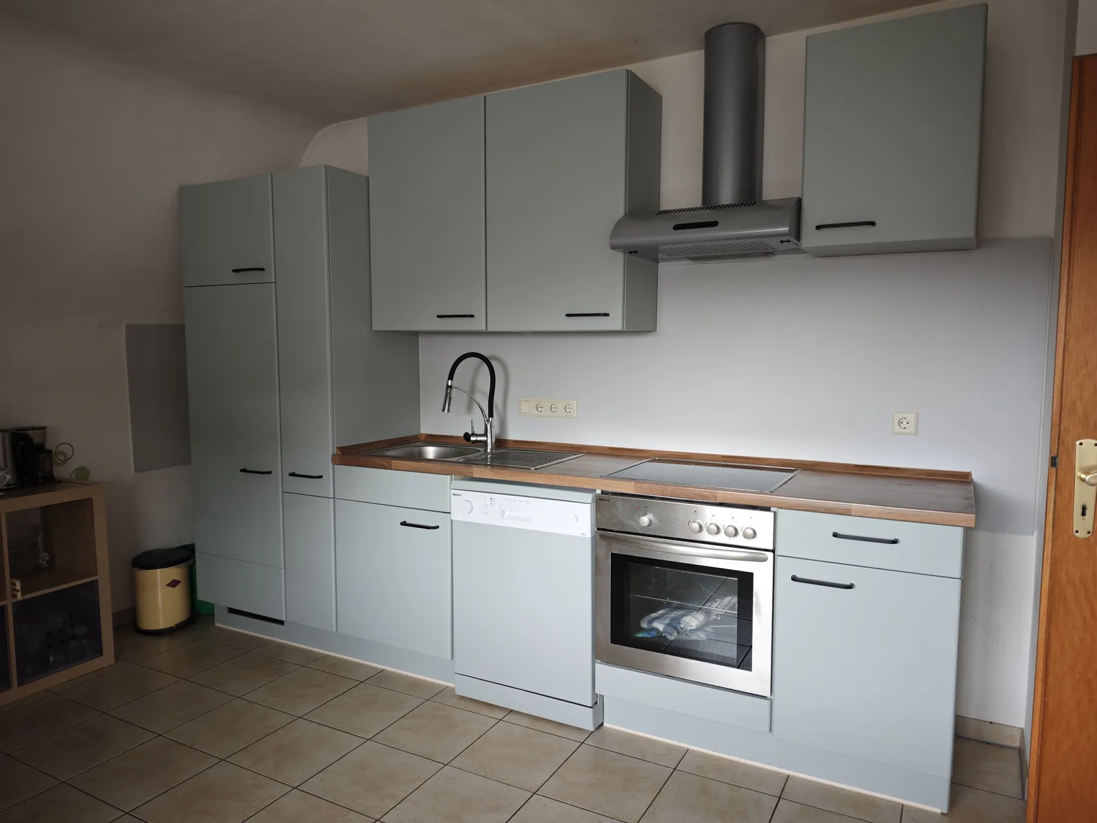Kleine Küchenzeile mit Fronten in mattem Salbeigrau, schwarzen Griffen, Arbeitsplatte in Holzoptik, Spüle, Geschirrspüler und Backofen in einer Reihe
