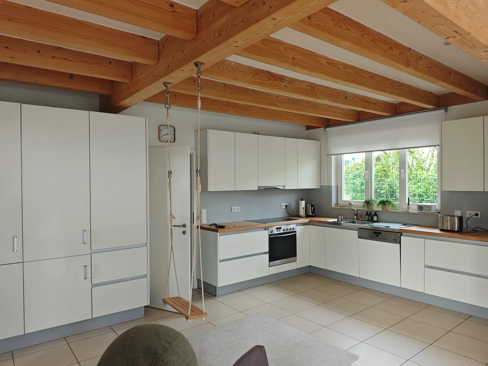 Mittlere L-Küche mit weißen grifflosen Fronten und grauem Sockel, Arbeitsplatte in Holzoptik, Hochschrankzeile links, Holzbalkendecke darüber