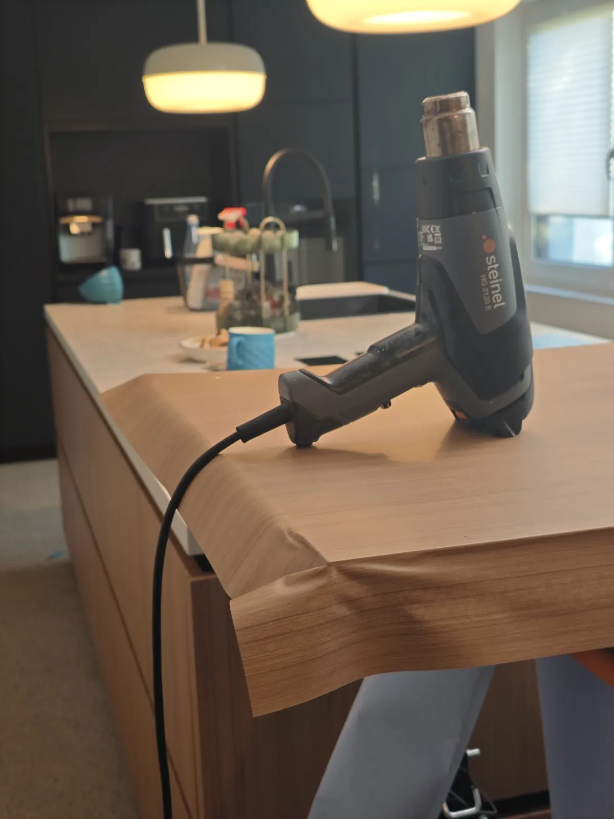 Nahaufnahme beim Folieren: Eine Heißluftpistole liegt auf der Arbeitsfläche, die Möbelfolie in Holzoptik wird mit Wärme um die Kante der Kochinsel gelegt