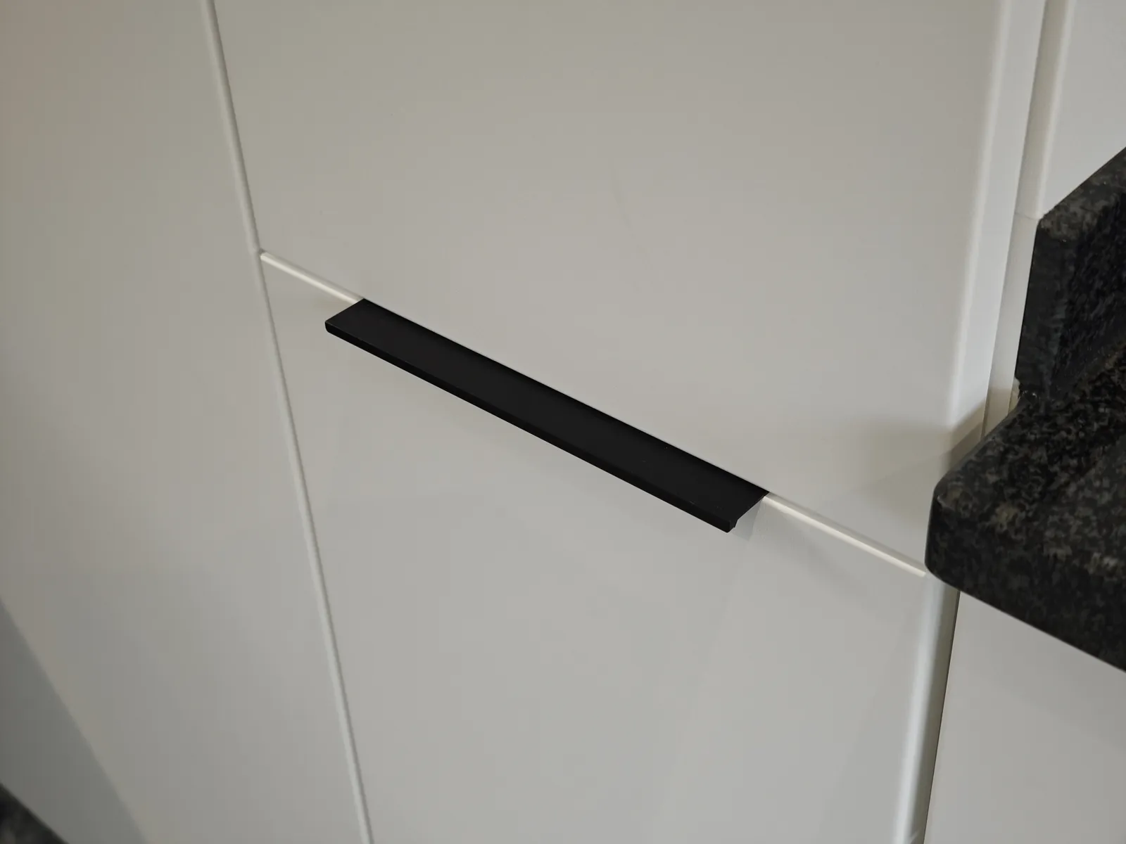 Detail einer weiß matt folierten Küchenfront mit neu montiertem schwarzem Profilgriff neben einer dunklen Steinarbeitsplatte